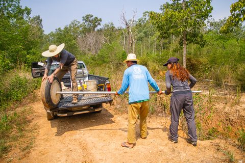 Three people carrying a steel roll-pole from a pickup truck toward a farm site