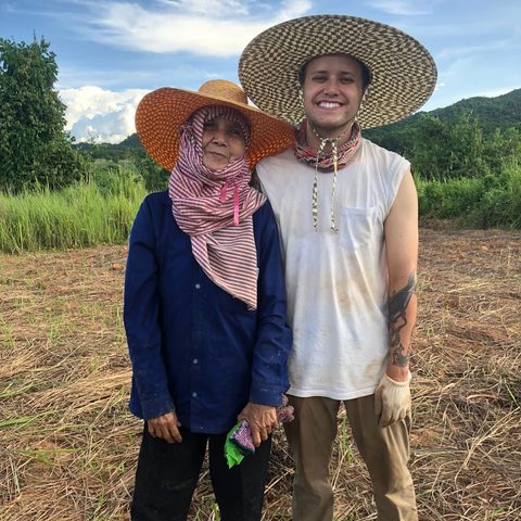 Tyler Nuckols standing with a farmer in a harvested field beside Kuiburi National Park, both in wide straw hats